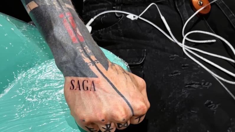 Machine Gun Kelly's right hand showing the new SAGA lettering tattoo near the edge of his blackout ink