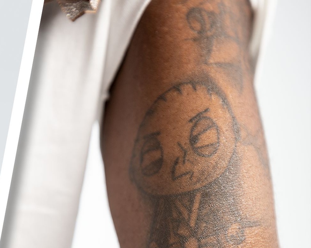 Burna Boy tattoos: Wide tattoo-tour crop showing the arm and hand lettering together